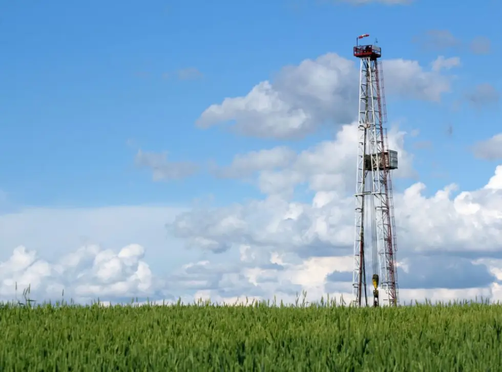A tall drilling rig stands above a green wheat field under a blue sky with fluffy clouds, indicating an active oil or gas exploration site