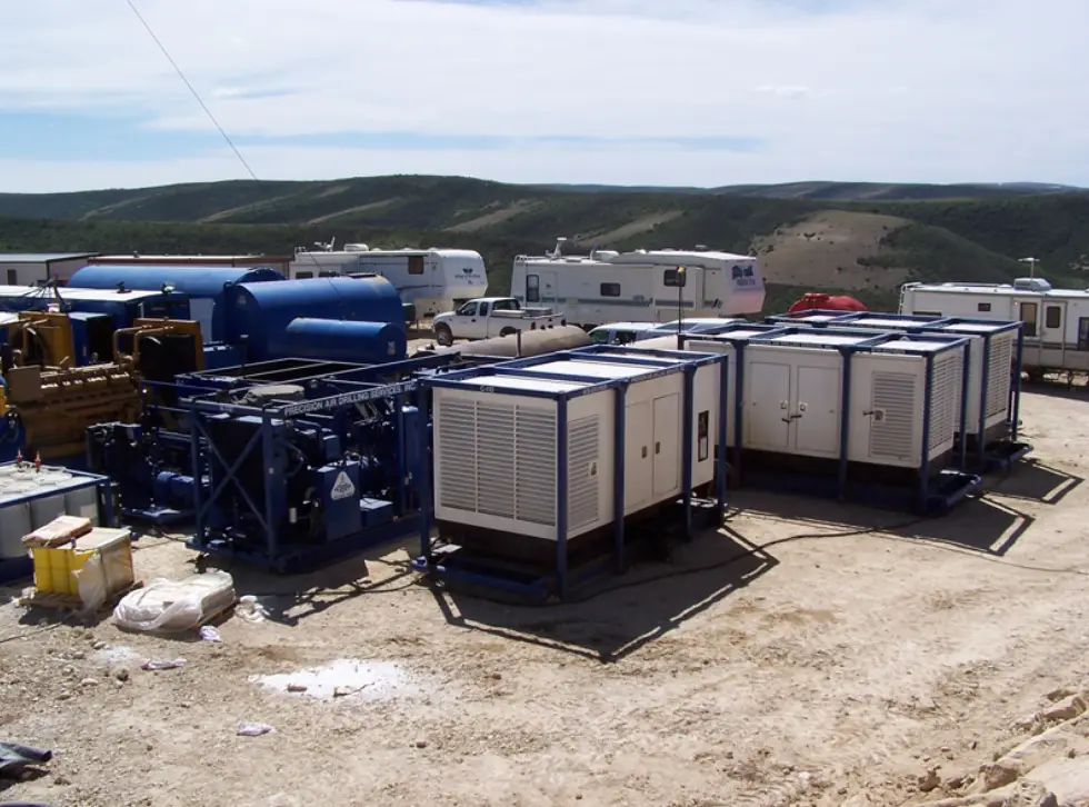 Industrial equipment is set up on a rocky terrain, with blue and white machinery surrounded by parked vehicles and rolling hills in the background
